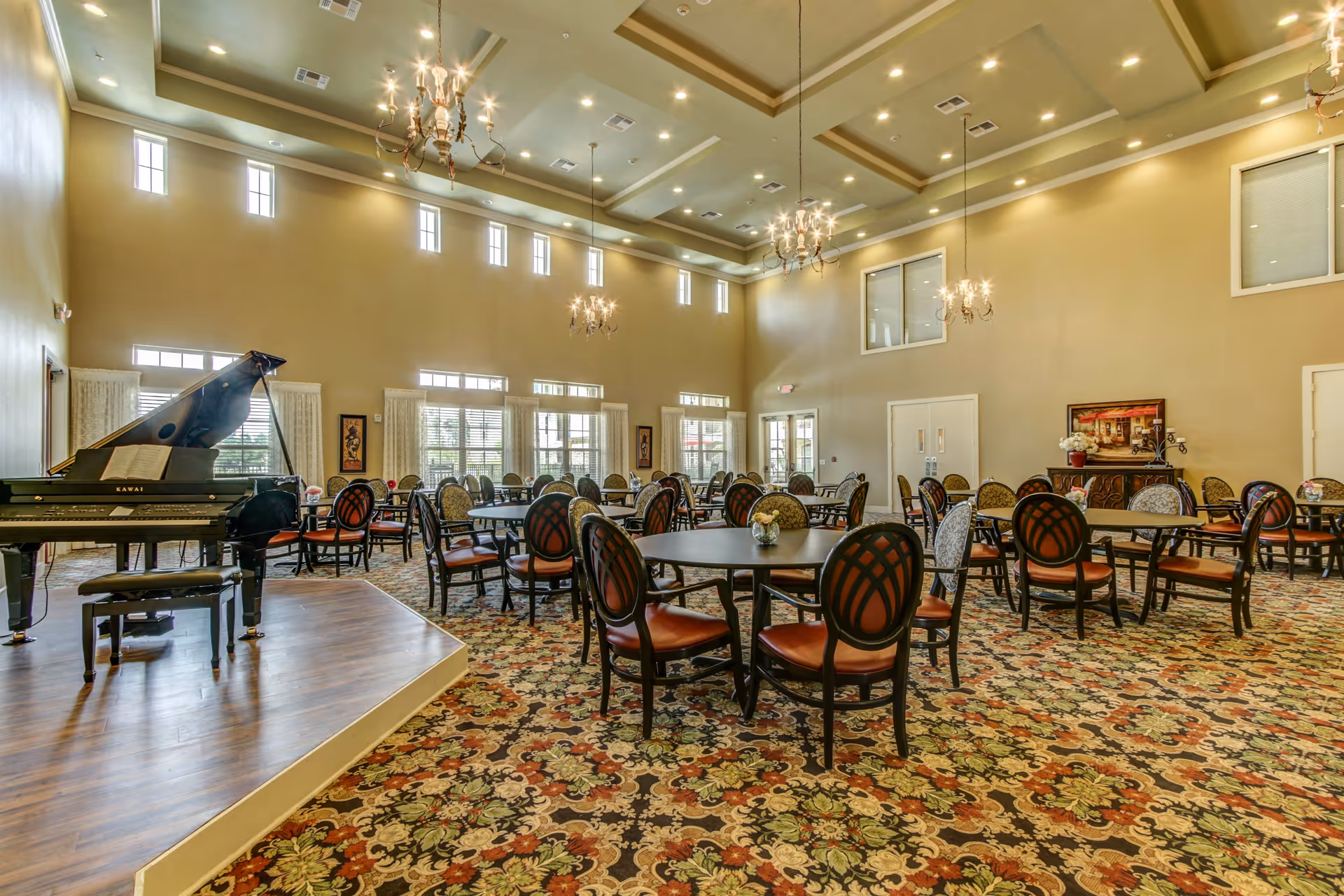 A spacious dining room with multiple round tables and chairs arranged neatly on a patterned carpet. The room features high ceilings with chandeliers, large windows allowing natural light, and a grand piano on a small stage to the left.