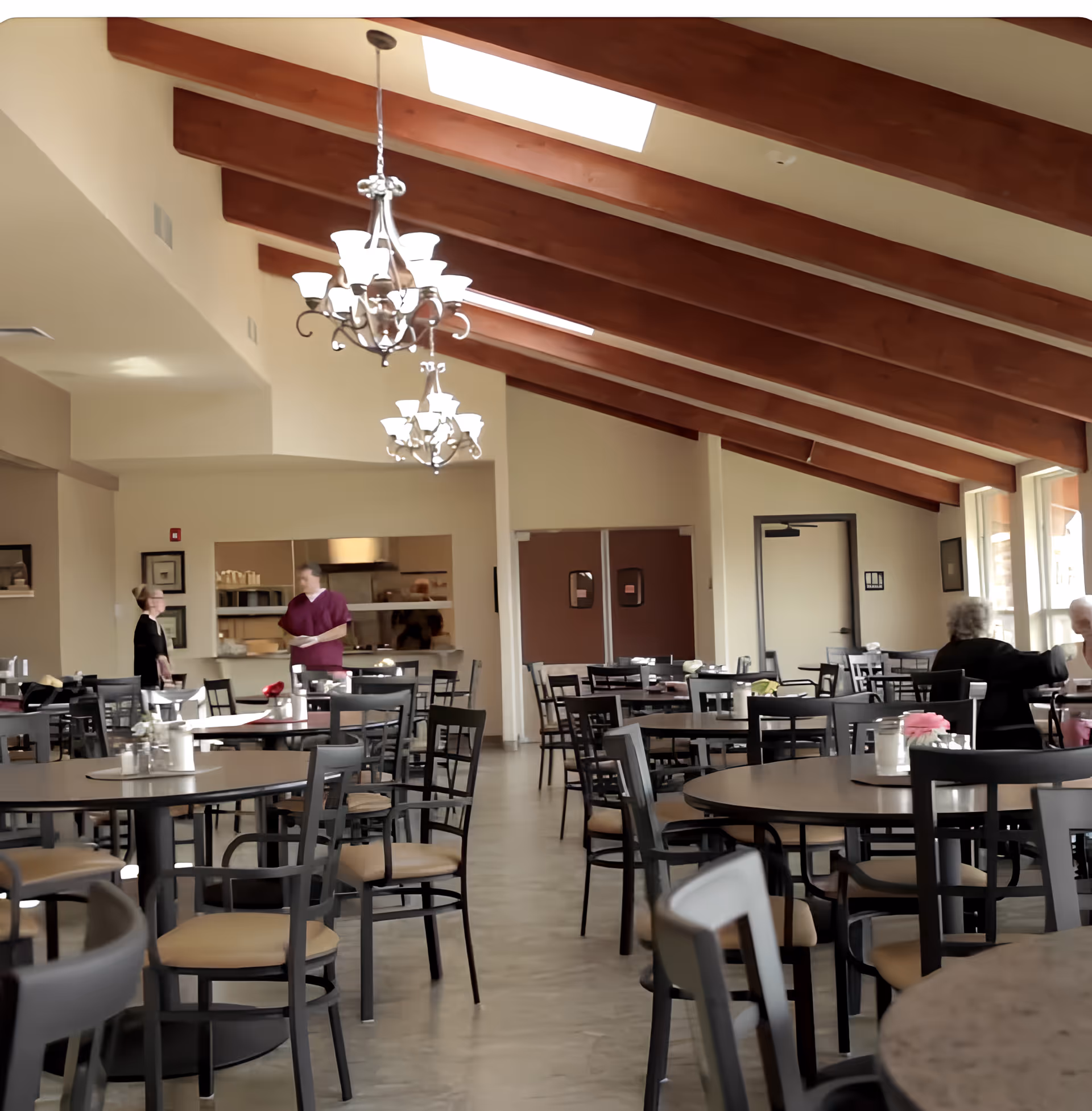 Spacious dining room with round tables and chairs under exposed wooden beams and chandeliers.