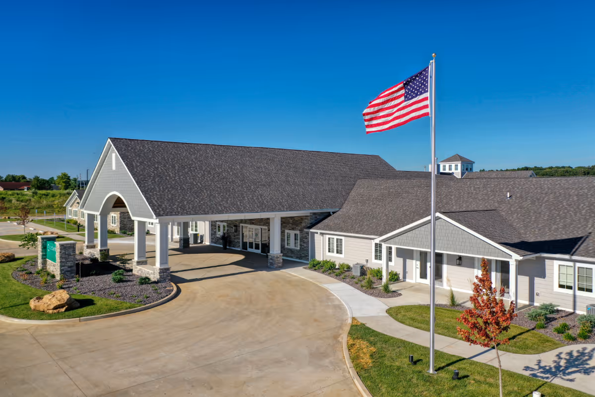 Exterior view of Cedarhurst Senior Living of Farmington building with a large covered entrance, an American flag on a flagpole, landscaped greenery, and a clear blue sky.