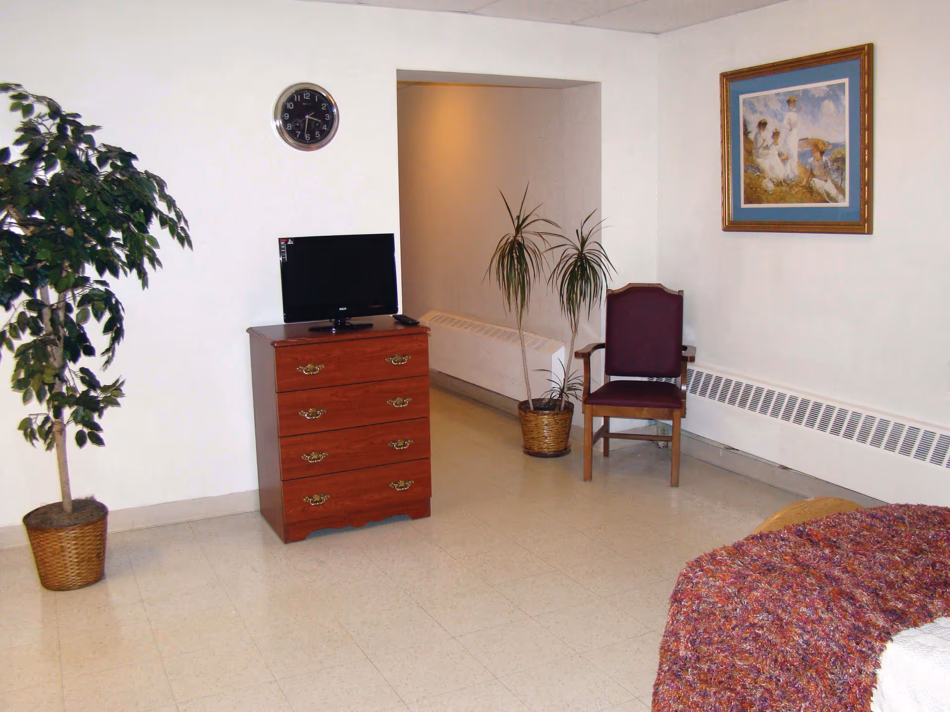 Interior nursing center room with a dresser holding a small TV, a chair, potted plants, wall clock and framed art, and a bed partially visible in the foreground.
