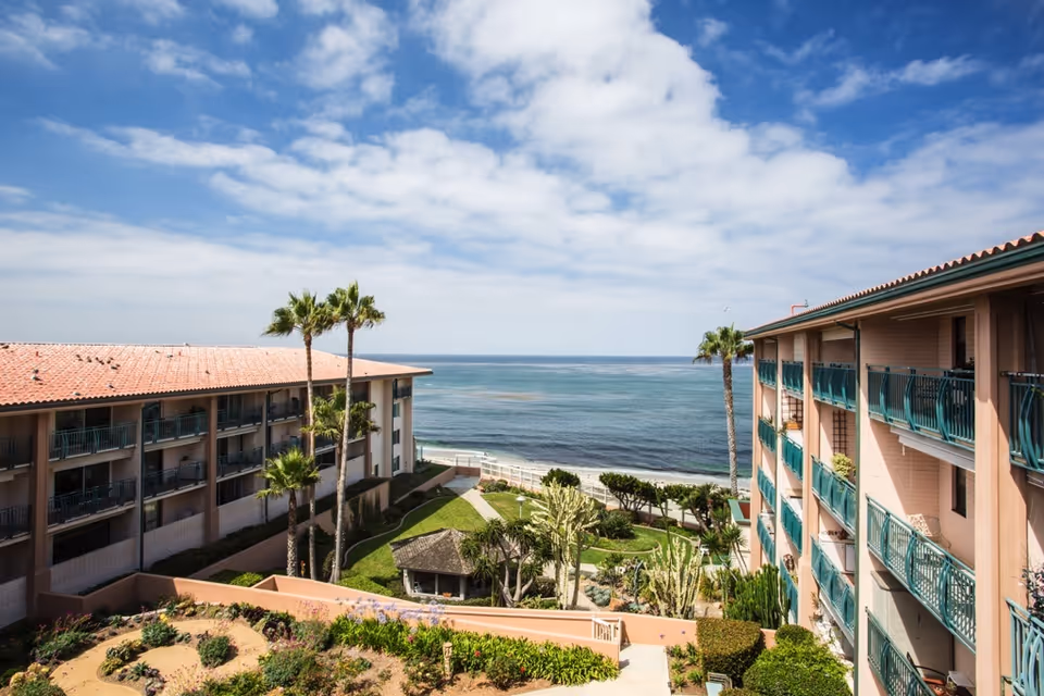 View of a senior living facility with two multi-story buildings facing each other, a landscaped garden with palm trees and shrubs in the center, and the ocean visible in the background under a partly cloudy sky.