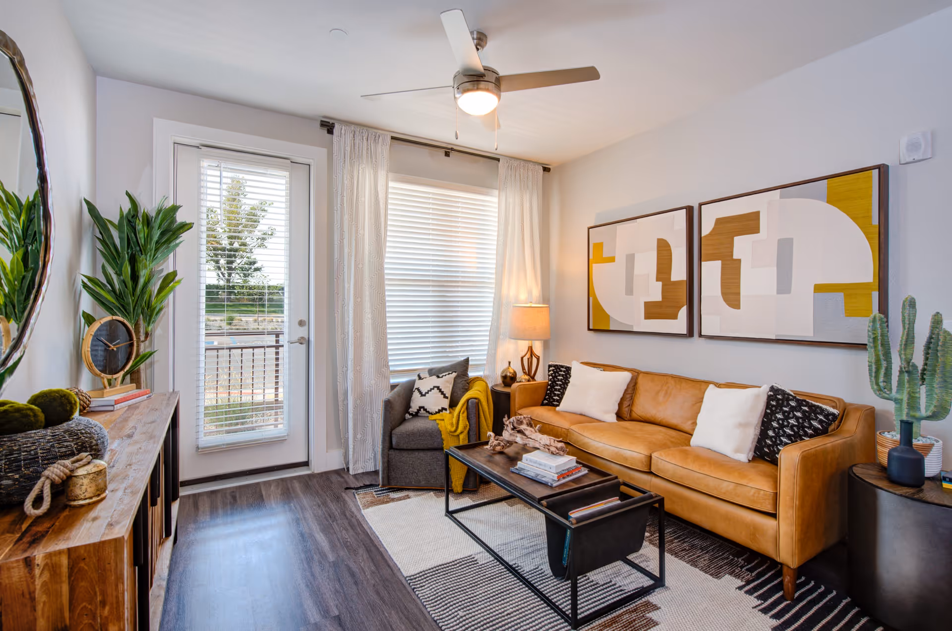 Well-lit contemporary living room with a tan leather sofa, armchair, coffee table, plants, wall art, and a door to a balcony.