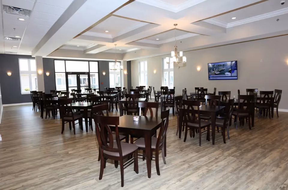 Spacious dining room with multiple dark wood tables and chairs, large windows, chandeliers, and a wall-mounted TV.