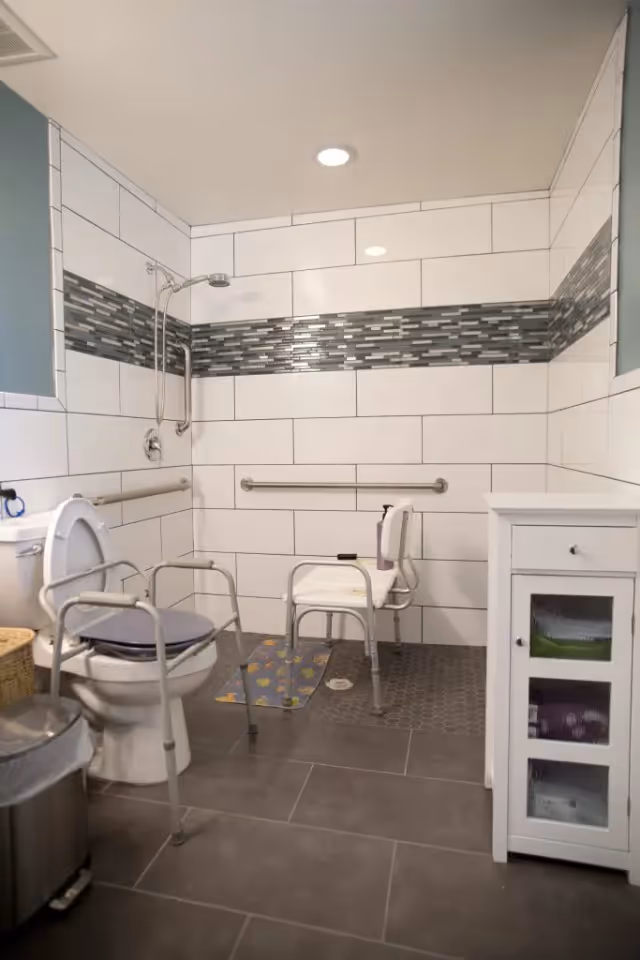 A bathroom designed for accessibility with a toilet equipped with safety rails, a shower area with a shower chair and grab bars, white tiled walls with a decorative horizontal strip of smaller tiles, and a white cabinet with frosted glass doors.