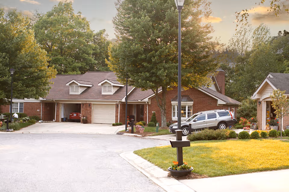 A residential street view in a senior living community showing brick houses with garages, a silver SUV parked in a driveway, green trees, a street lamp, and a mailbox with flowers planted at its base.