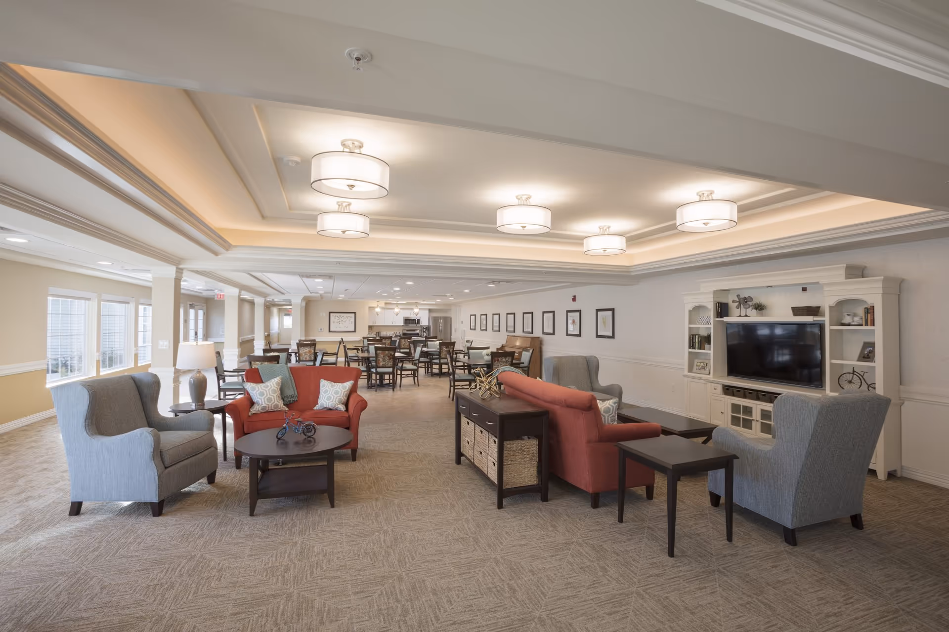 A spacious and well-lit assisted living common area featuring comfortable seating including red sofas and gray armchairs arranged around coffee tables. The room has a large TV on a white entertainment center, multiple ceiling lights, and a dining area with tables and chairs in the background. Large windows on the left side allow natural light to fill the space.