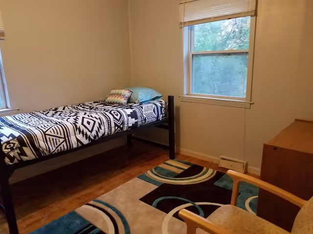 A small bedroom with a single bed featuring a black and white patterned bedspread and two pillows. There is a window with a blind partially open, a wooden dresser, a chair with a light-colored cushion, and a colorful area rug on the wooden floor.