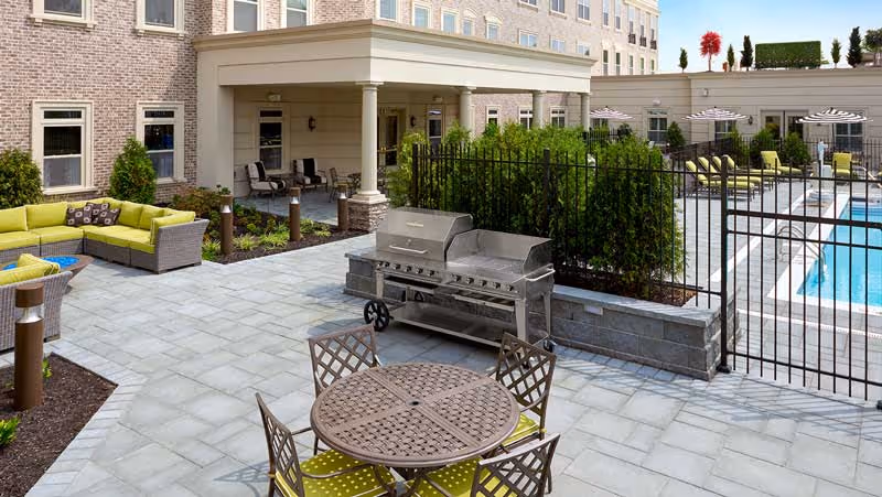 Outdoor patio area at a senior living facility featuring a round metal table with four chairs, a large stainless steel barbecue grill, a green cushioned sectional sofa, and a fenced swimming pool with lounge chairs and umbrellas. The building exterior is visible in the background with multiple windows and a covered seating area.