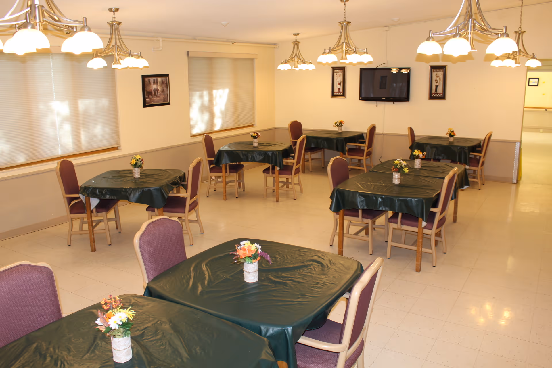 A dining room with multiple tables covered in dark green tablecloths, each table having a small flower arrangement centerpiece. The room has beige walls, windows with blinds, several ceiling light fixtures, and a wall-mounted television.