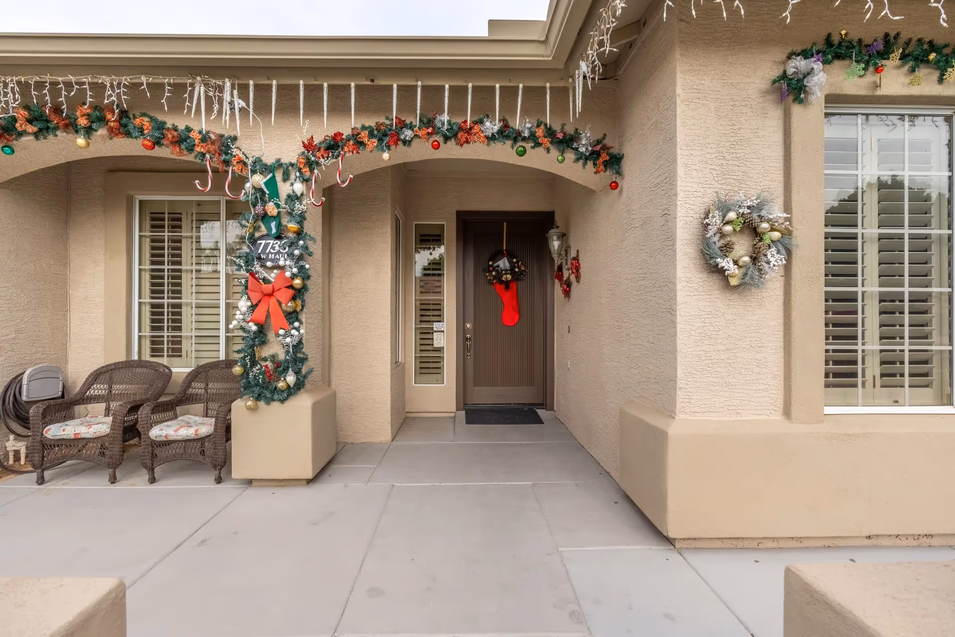 Front entrance of a beige stucco building decorated with Christmas garlands, wreaths, and icicle lights. There are two wicker chairs with cushions on the left side under an archway, and a red Christmas stocking hanging on the front door. The address number 7735 is displayed on a decorated pillar near the door.