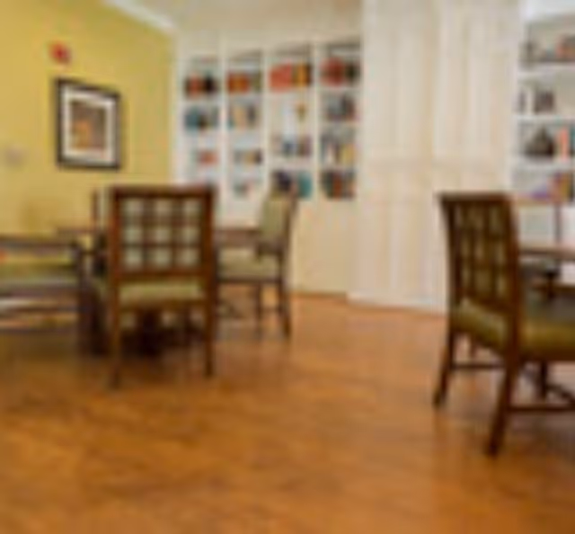 A cozy interior room with wooden flooring, several wooden chairs with green cushions around tables, and white built-in shelves filled with books and decorative items against a light yellow wall.