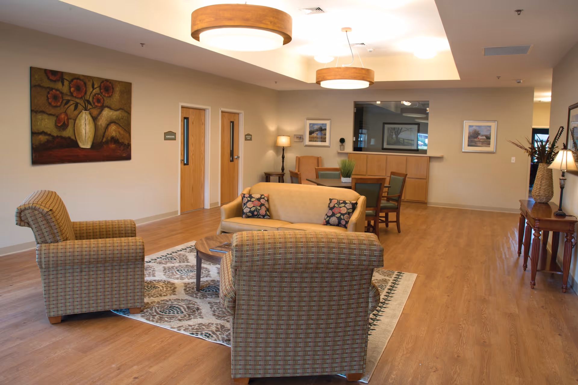 A spacious and well-lit common area in Maplewood Healthcare Center featuring a seating arrangement with a beige loveseat and two patterned armchairs around a wooden coffee table on a decorative rug. The room has wooden flooring, neutral-colored walls, framed artwork, a wooden console table with a lamp and decorative items, and two wooden doors labeled 'Conference'. Ceiling lights with wooden accents illuminate the space.