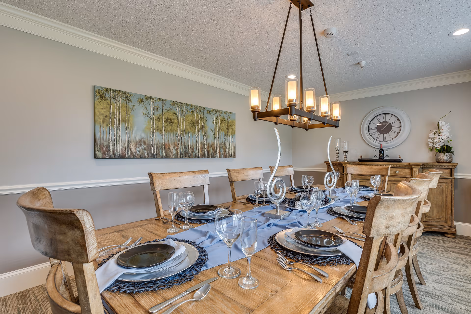 A dining room with a wooden table set for eight people, featuring plates, glasses, silverware, and decorative candle holders. The room has a large rectangular chandelier hanging above the table, a sideboard with a clock and flower arrangement, and a painting of trees on the wall.