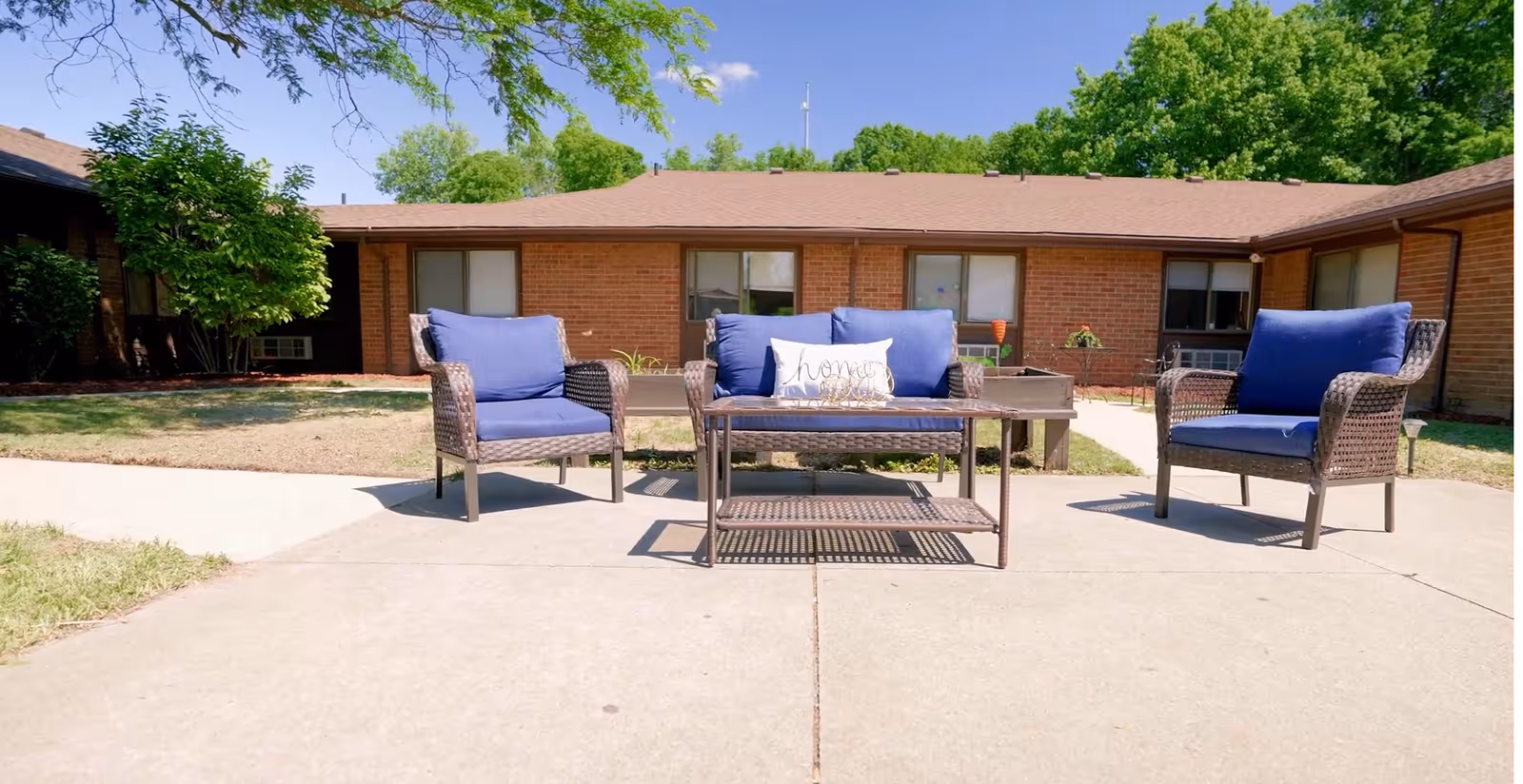 Outdoor patio area with three wicker chairs with blue cushions and a matching coffee table. The chairs and table are placed on a concrete surface in front of a single-story brick building with several windows. There are some green trees and bushes in the background under a clear blue sky.
