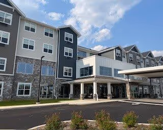 Exterior view of a modern multi-story senior living facility building with a covered entrance, stone and siding facade, and a clear blue sky with some clouds.