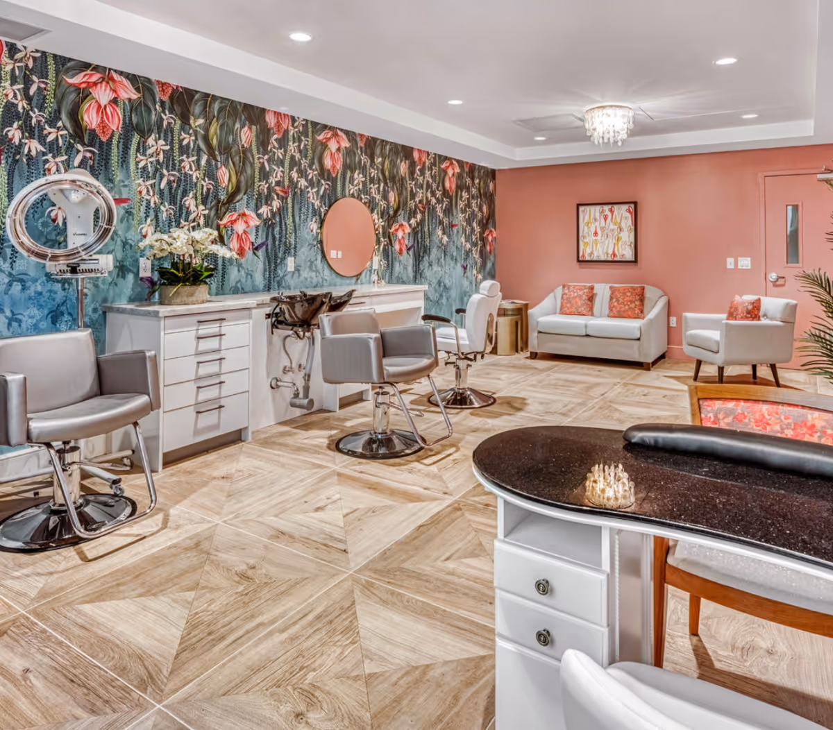 A bright and stylish salon area in a senior living facility featuring three salon chairs with mirrors and hair washing stations along a floral patterned wall. The room has a light wood patterned floor, a white manicure table with a black countertop in the foreground, and a cozy seating area with a white loveseat and armchair with coral cushions against a coral-colored wall.