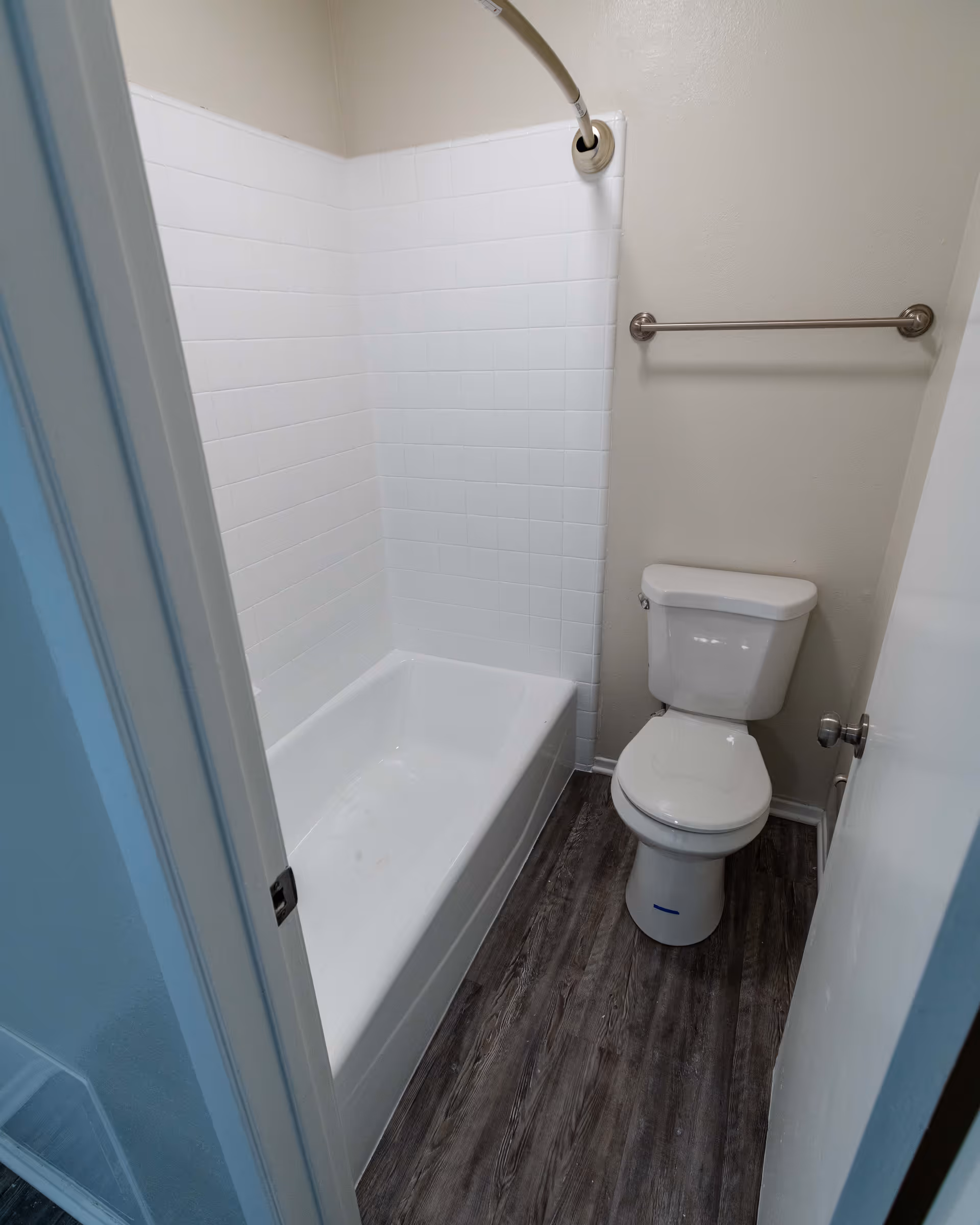 Small bathroom with a white bathtub and tiled walls, a white toilet, a towel bar on the wall, and wood-patterned flooring.