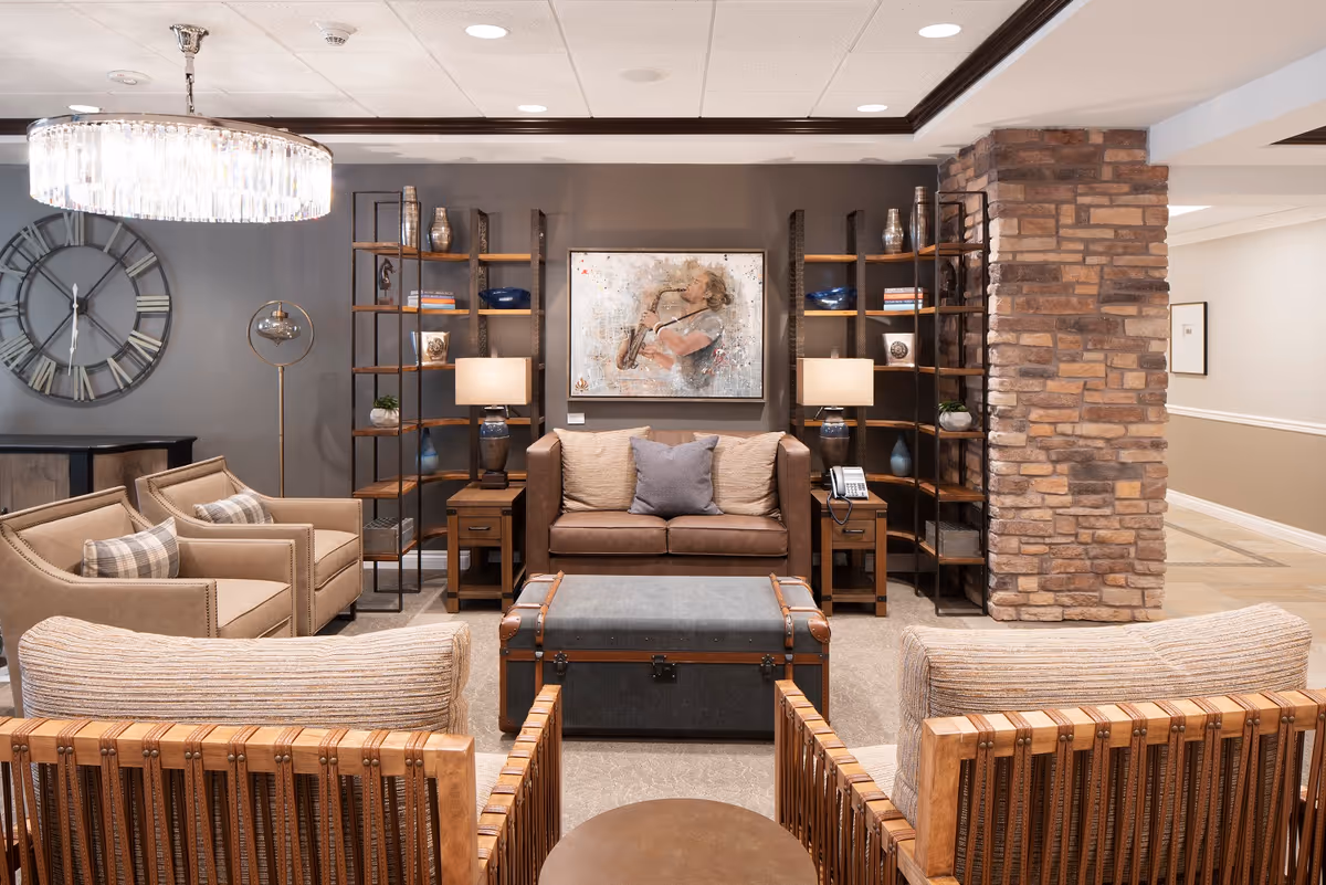 A cozy senior living room with a brown leather sofa, two beige armchairs with plaid pillows, and two wooden chairs with cushions. The room features a large wall clock, two tall bookshelves with decorative items, a painting of a woman playing a saxophone, two table lamps, a floor lamp, and a trunk-style coffee table. The walls are painted gray with a stone pillar on the right side.