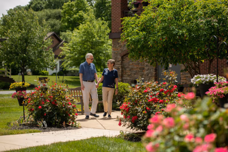 An elderly couple holding hands and walking along a paved path surrounded by lush greenery and vibrant flowers in a garden area near a brick building.