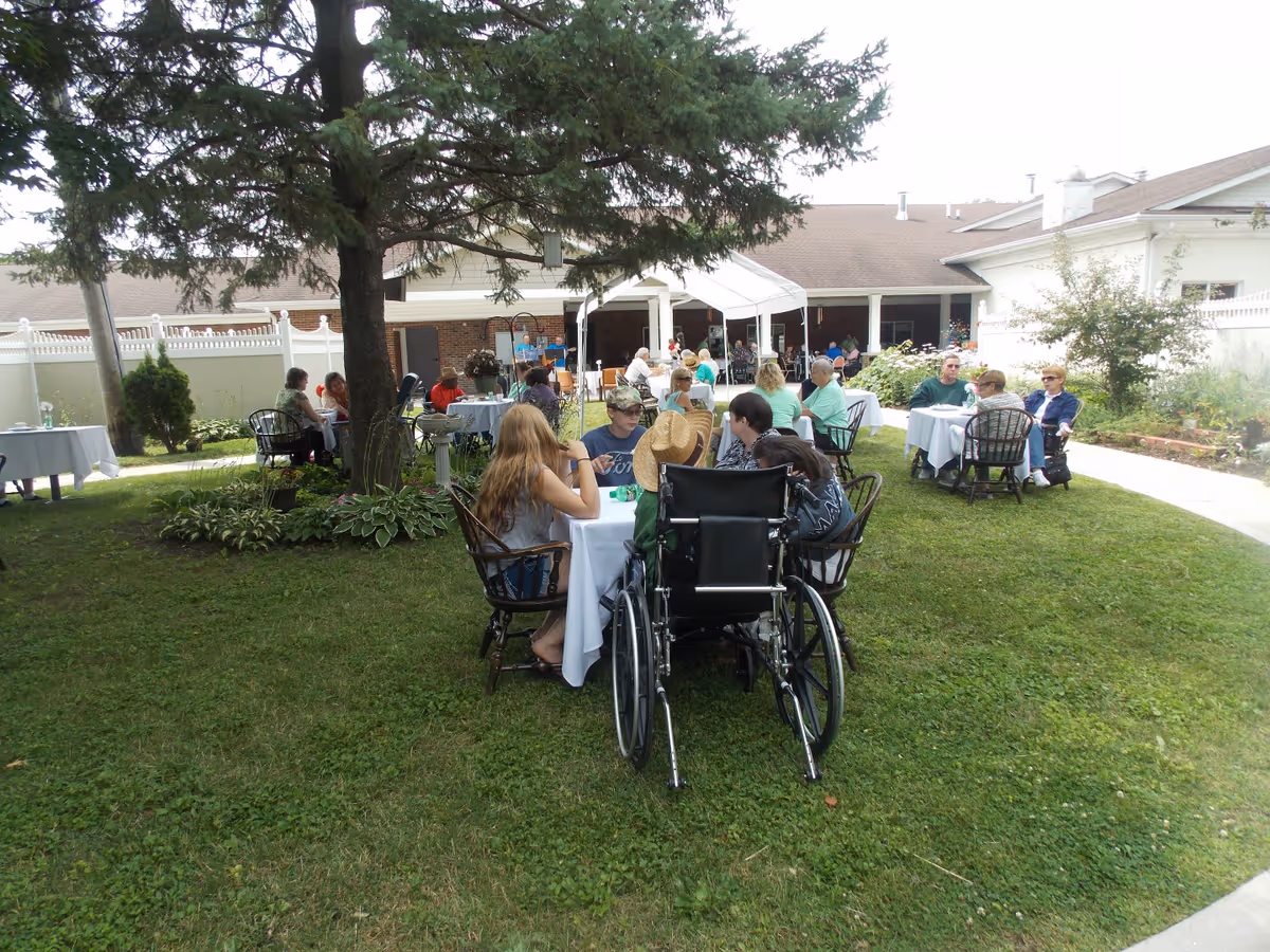 People sitting at tables on a grassy courtyard of a memory care residence with wheelchairs and the facility building visible.