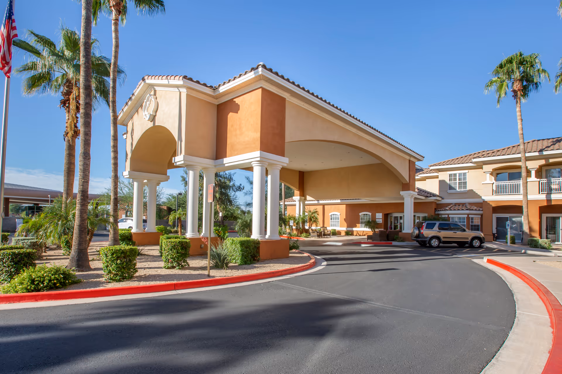 Front entrance of a Mediterranean-style senior living building with a covered porte-cochere, palm trees, and a curved driveway.