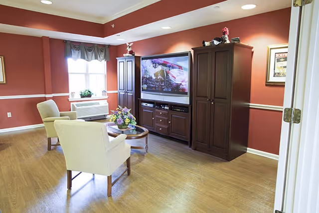 A cozy living room with two beige armchairs facing a large flat-screen TV mounted on a dark wooden entertainment center. The room has red walls with white trim, a window with a valance, a round wooden coffee table with a flower arrangement, and wood flooring.