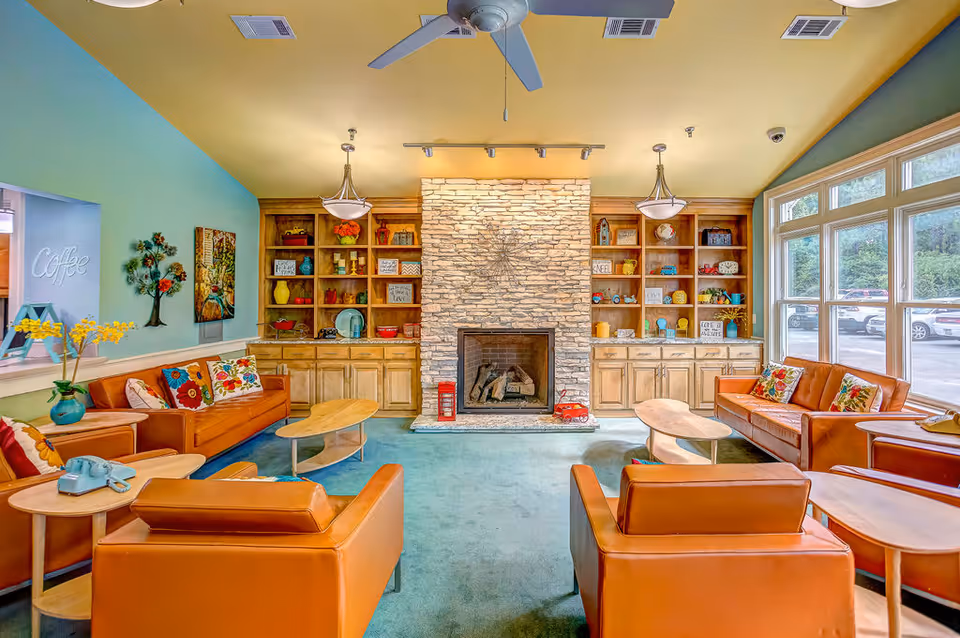 Bright communal lounge with orange leather sofas arranged around a stone fireplace and built-in shelving under large windows.