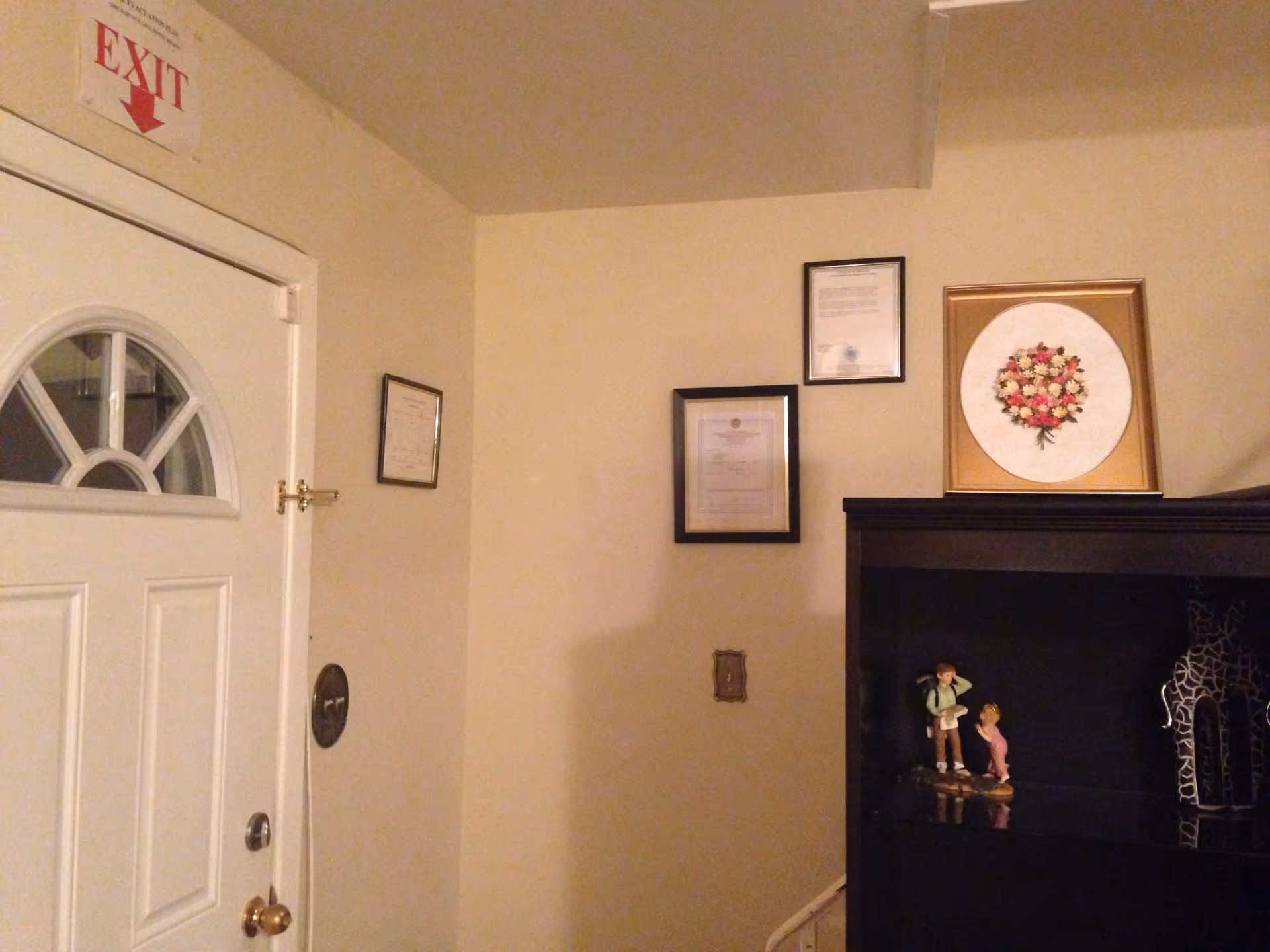 Interior corner of a room showing a white door with a small window and an exit sign above it. On the adjacent wall, there are three framed certificates or documents. A black cabinet with decorative items including a small figurine of two children and a giraffe sculpture is also visible. A framed floral artwork is placed on top of the cabinet.