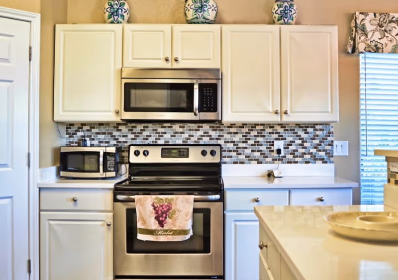 A modern kitchen featuring white cabinets, a stainless steel microwave mounted above a stainless steel electric stove with a grape-themed towel hanging on the oven handle. There is a small microwave on the countertop to the left, a mosaic tile backsplash in shades of brown, beige, and white, and decorative vases on top of the cabinets. A window with blinds and a floral valance is visible on the right side.