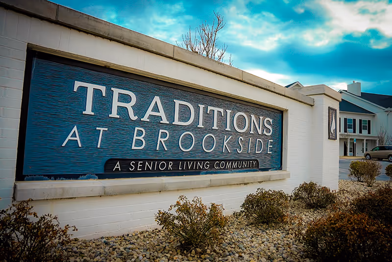 Entrance sign reading "TRADITIONS AT BROOKSIDE — A Senior Living Community" with landscaping and the facility building in the background.