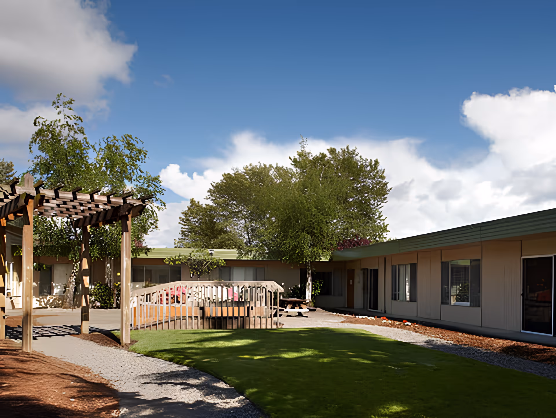 Outdoor courtyard area of a senior living facility with a wooden pergola, a small wooden bridge, green lawn, trees, and a building with large windows under a partly cloudy blue sky.