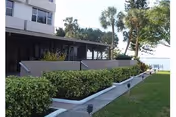 Exterior view of Presbyterian Villas of Tampa showing a building with a covered patio area, surrounded by greenery and palm trees, with a walkway and lawn leading towards a body of water in the background.