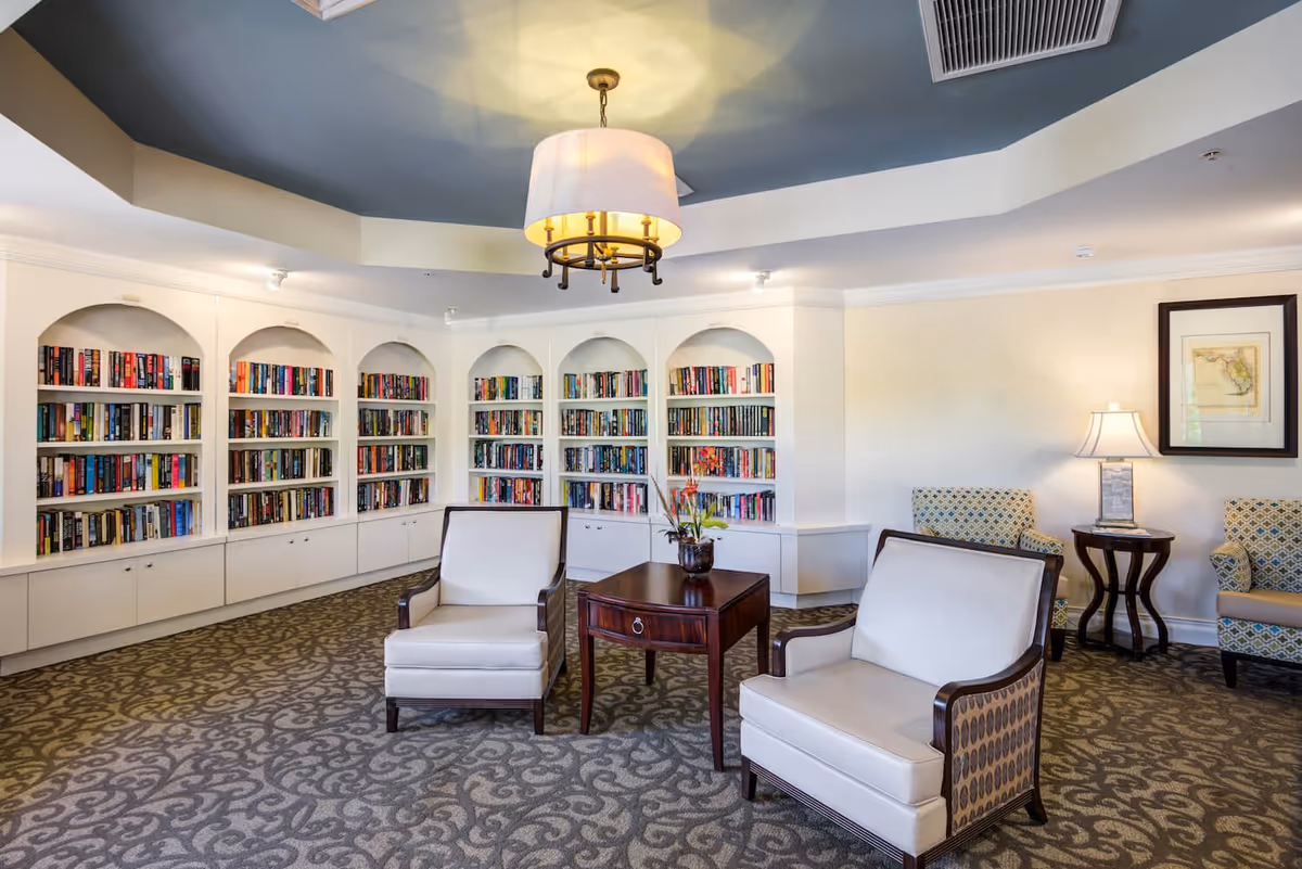 A cozy reading room with built-in white bookshelves filled with books along the back wall. Two cushioned armchairs with wooden frames are positioned around a small wooden table with a flower arrangement. The room has patterned carpet, a ceiling light fixture, and additional seating with patterned armchairs and a side table with a lamp.