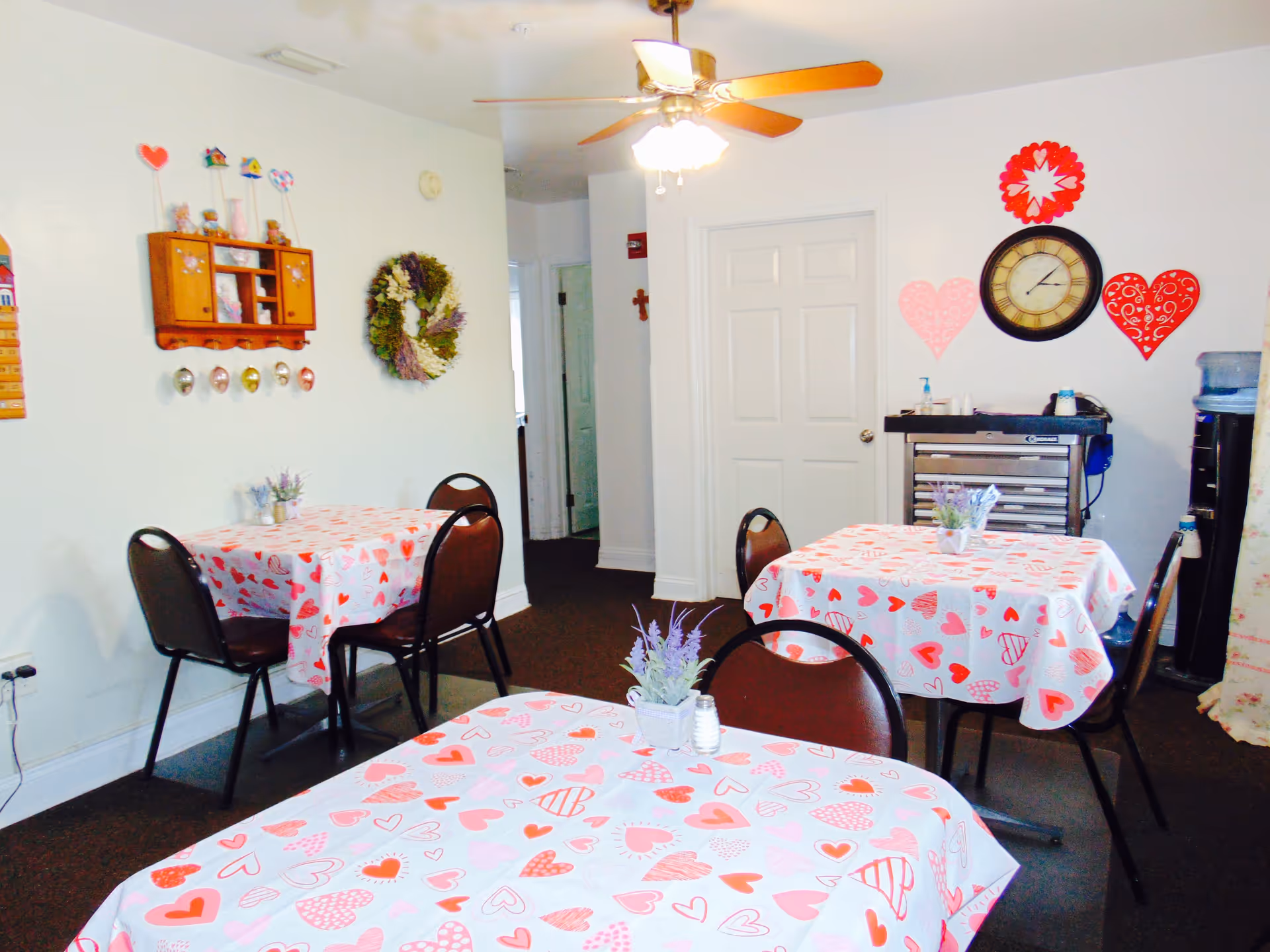 A small dining area with three tables covered in white tablecloths decorated with red and pink hearts. Each table has a small potted plant centerpiece. The room has white walls adorned with heart-shaped decorations, a clock, a wreath, and a small wooden shelf with decorative items. There are several brown chairs around the tables, a ceiling fan with lights, and a water dispenser in the corner.