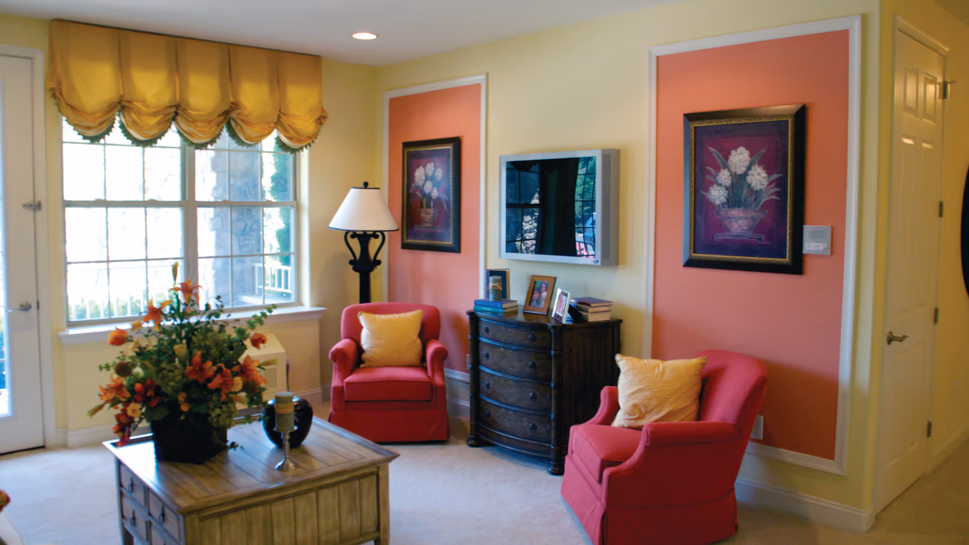 A cozy living room area with two red armchairs each with a yellow pillow, a wooden chest of drawers with books and framed photos on top, a wall-mounted flat-screen TV, two framed floral paintings on a peach-colored accent wall, a large window with yellow valance curtains, a wooden coffee table with a floral arrangement and decorative items, and a floor lamp in the corner.