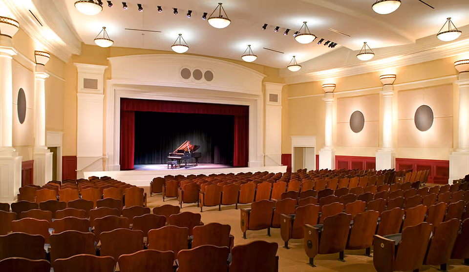 Empty auditorium with rows of wooden seats facing a stage with a grand piano and red curtains under warm lighting.