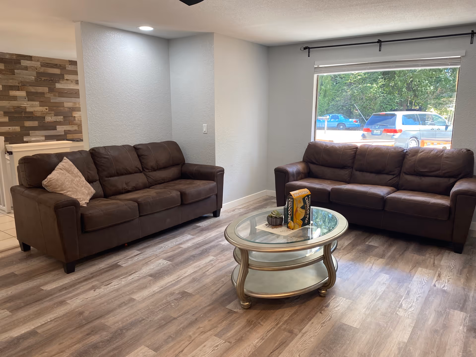 A living room with two brown leather sofas facing each other, a glass coffee table with a small plant and a box on it, wood-style flooring, and a large window showing parked cars and greenery outside.