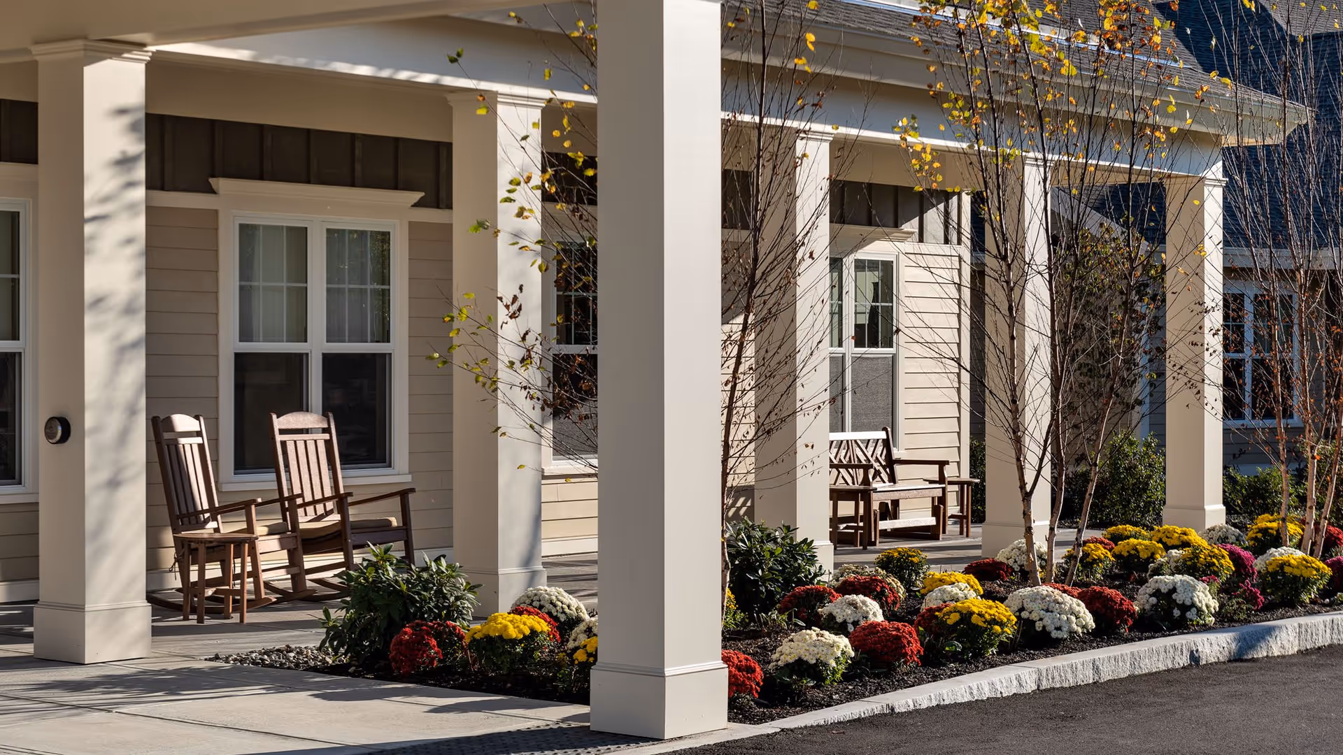 Outdoor covered porch area at a senior living facility with wooden rocking chairs and benches. The porch is supported by large white columns and bordered by a flower bed with colorful red, yellow, and white flowers. Leafless trees are planted along the flower bed, and the building exterior features beige siding and white-framed windows.