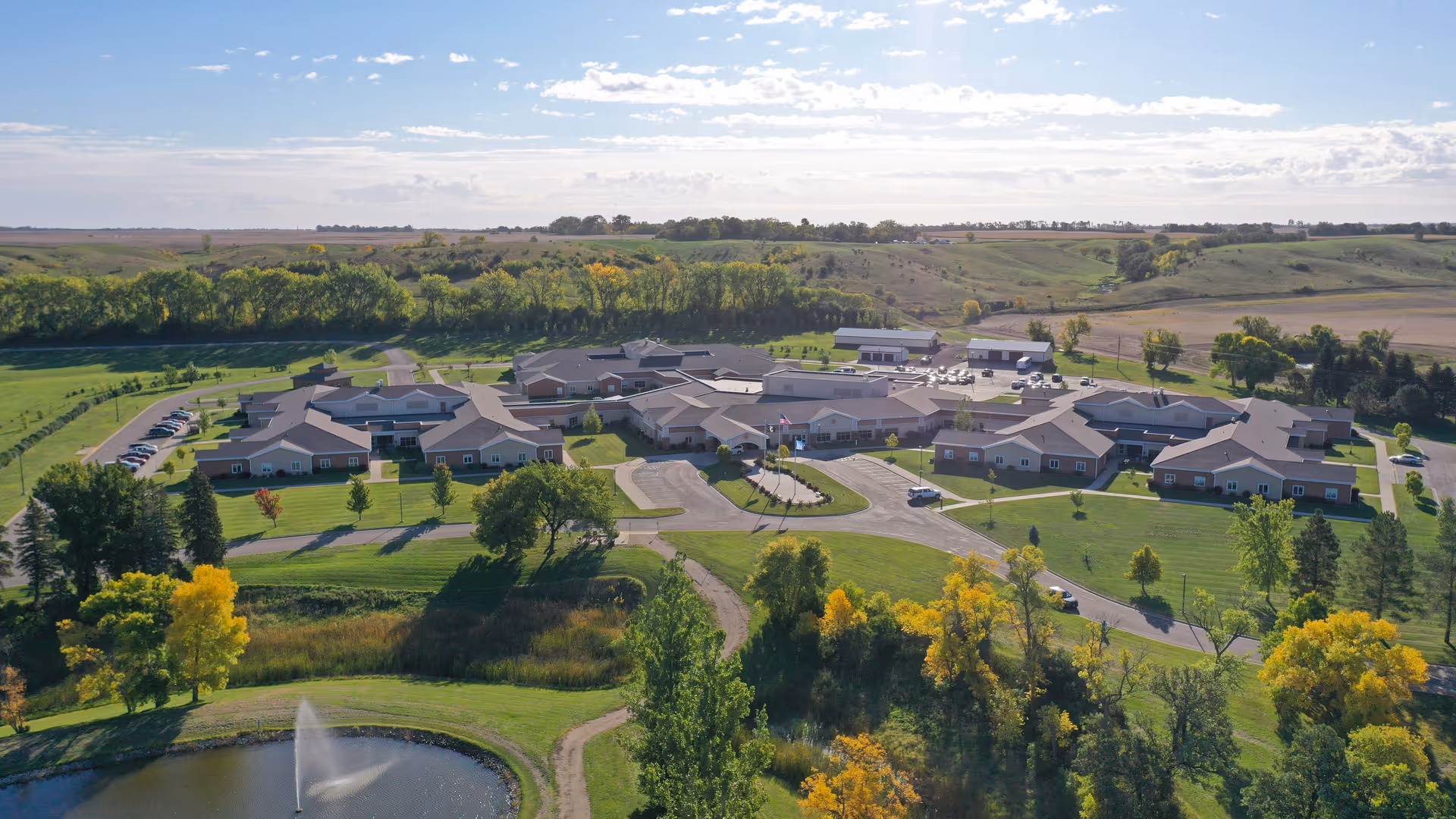 Aerial view of the North Dakota Veterans Home facility surrounded by green lawns, trees, and open fields under a partly cloudy sky. The complex consists of multiple connected buildings with parking areas and a small pond with a fountain in the foreground.