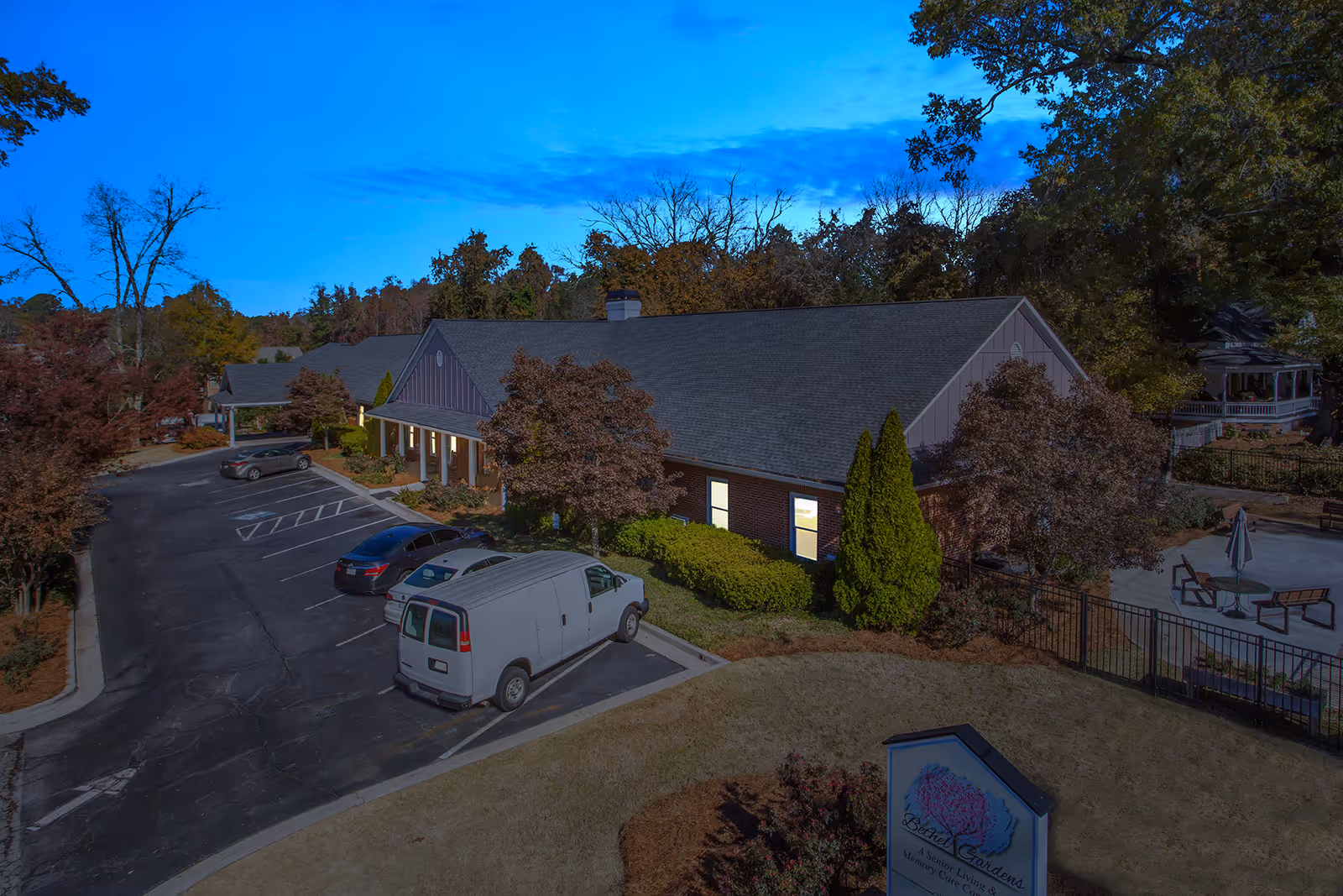Exterior view of Bethel Gardens Assisted & Memory Care facility during dusk, showing a single-story building with a parking lot in front, several parked vehicles, trees with autumn foliage, and a fenced outdoor seating area with tables and umbrellas.