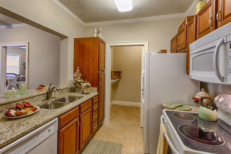 Well-appointed kitchen with granite countertops, double sink, wooden cabinets, white appliances and an open pantry doorway.