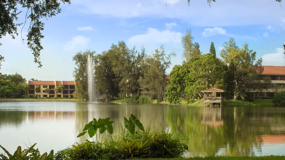 A serene outdoor scene at American House Sarasota featuring a large pond with a water fountain in the center, surrounded by lush greenery and trees. There is a small gazebo on the right side near the water's edge, and buildings with red-tiled roofs are visible in the background under a partly cloudy blue sky.