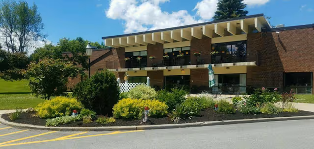 Exterior view of a single-story brick building with a flat roof and large windows. The building is surrounded by a well-maintained garden with various shrubs and flowers. A paved driveway and parking area are visible in the foreground under a partly cloudy sky.