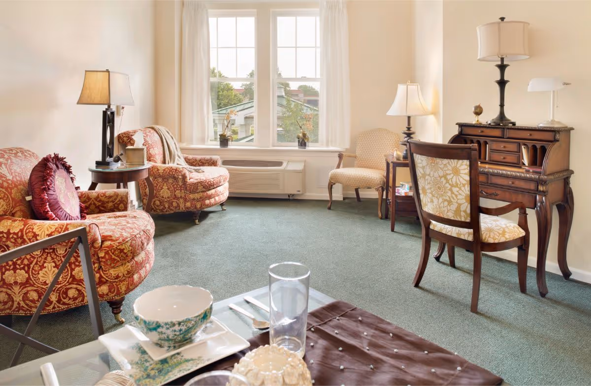 A cozy living room with two patterned armchairs, a wooden desk with a matching chair, two table lamps, and a window with white curtains letting in natural light. A glass coffee table with dishes and a glass is in the foreground.