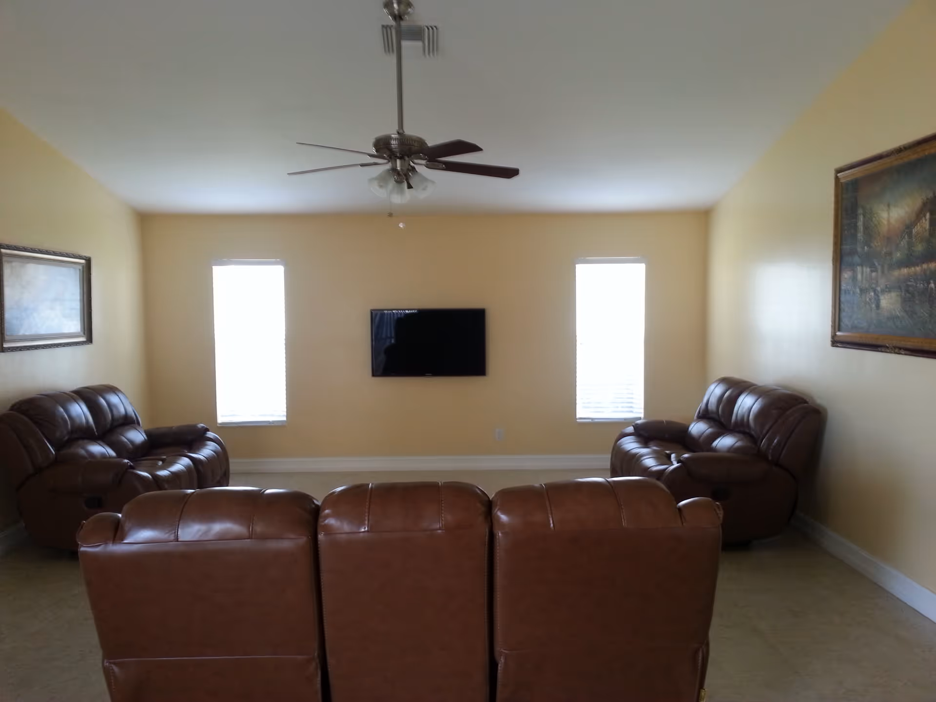 A living room with three brown leather recliners facing a wall-mounted flat screen TV. The room has two narrow windows on either side of the TV, a ceiling fan with lights, and two framed paintings on the walls. The walls are painted a light yellow color.