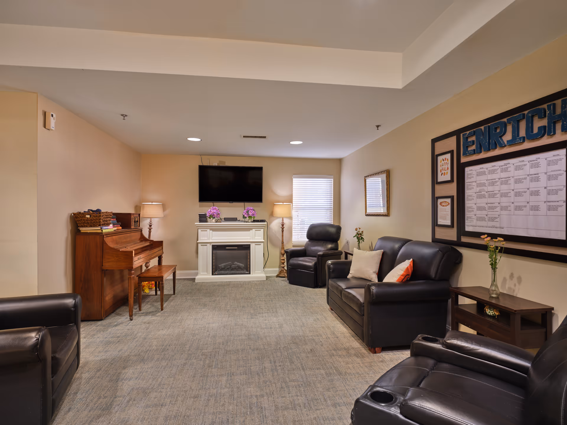 A cozy living room area in an assisted living facility featuring a wooden piano with a bench on the left, a white electric fireplace with a TV mounted above it in the center, and several leather chairs and a sofa arranged around the room. The walls are beige, and there is a large bulletin board labeled 'ENRICH' on the right wall. The room is softly lit with lamps and natural light coming through a window with blinds.