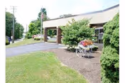 Exterior view of a care facility entrance with greenery, a decorative cart, and a paved driveway.