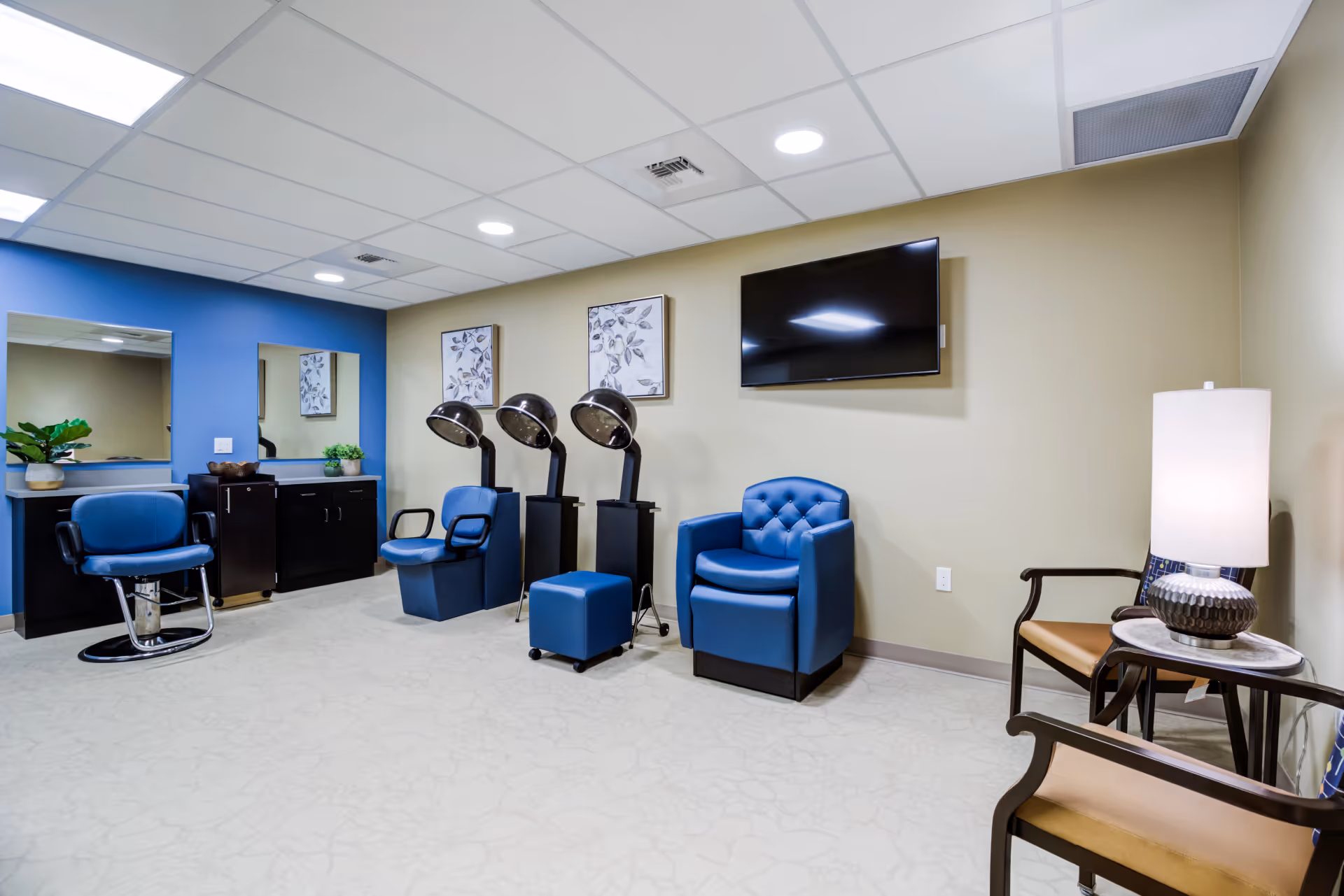 Interior view of a senior living community salon area with blue salon chairs, hair dryers, a blue armchair, two wooden chairs with tan cushions, a side table with a lamp, two framed botanical prints on the wall, and a wall-mounted flat screen TV.