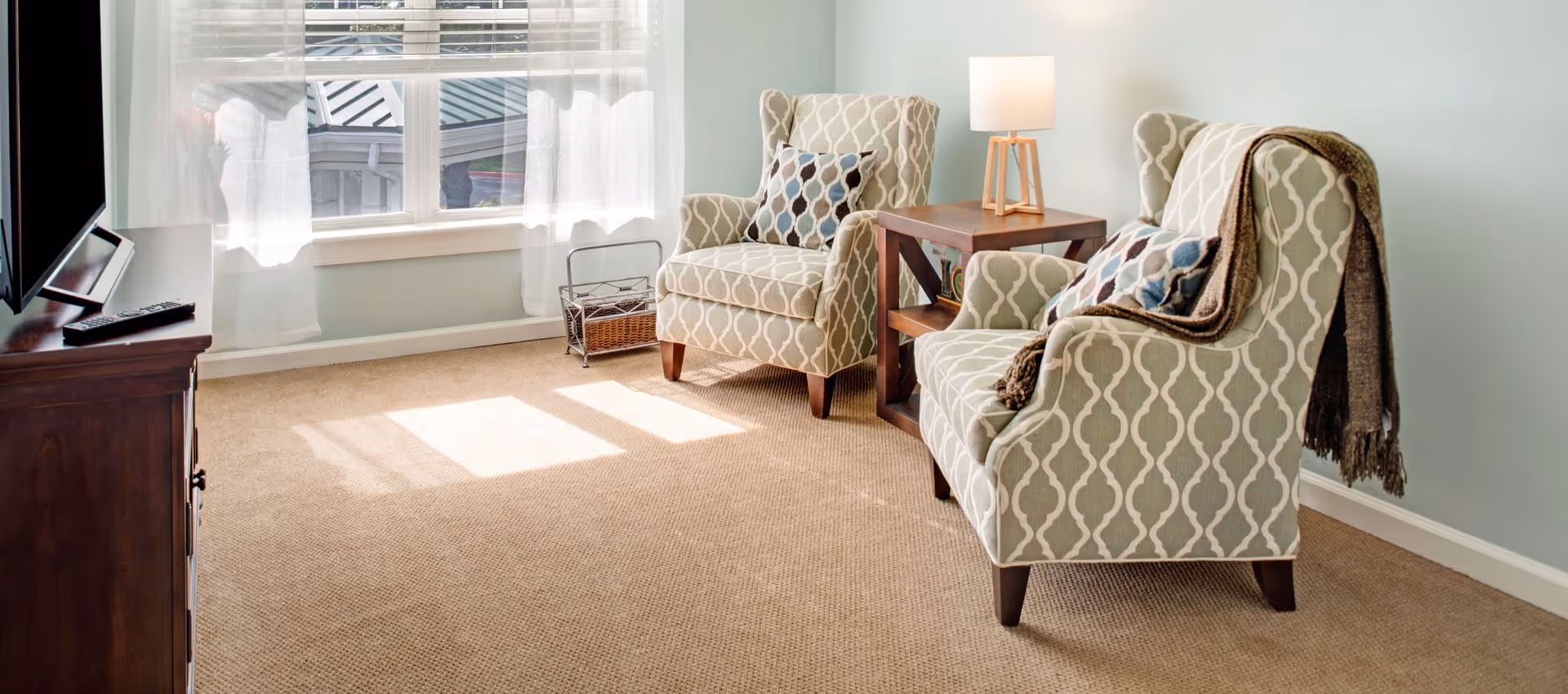 A cozy living room area with two patterned armchairs, each with a decorative pillow. A brown throw blanket is draped over one chair. Between the chairs is a wooden side table with a modern lamp. Sunlight streams through a window with sheer white curtains, illuminating the beige carpeted floor. A wooden TV stand with a television and remote controls is visible on the left side.