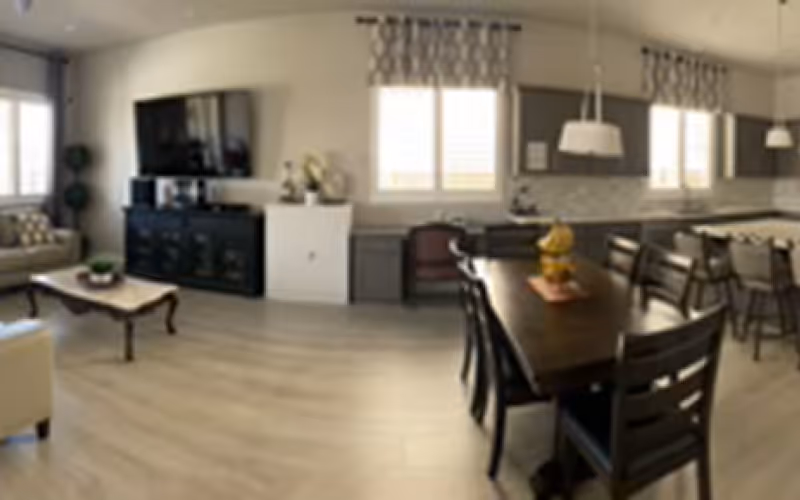 Open-plan living and dining area with a wooden dining table and chairs, sofa, television, and windows.